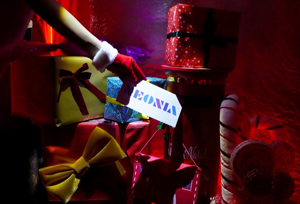 Mão com luva de Natal a segurar um voucher Neonia entre presentes coloridos, na sala temática natalícia do museu.