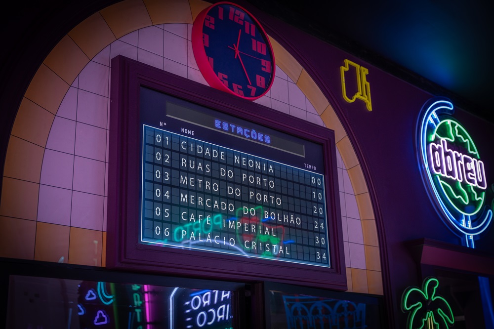 Painel de estações na sala Cidade Neonia que mostra os nomes das salas do museu imersivo e o tempo estimado para explorar cada uma.