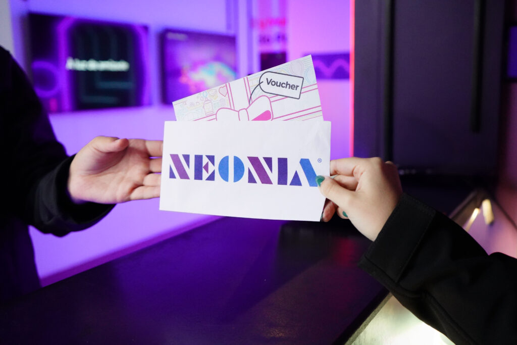 Two people exchanging a Neonia voucher, with the logo visible, in a space lit by colourful lights inside the immersive museum.